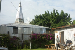 »Diesmal fuhr King Charles gleich zu einem Seiteneingang des Krankenhauses, der direkt zur Intensivstation führte. Daneben stand eine Kirche, zur Gänze aus Wellblechplatten errichtet, selbst der spitz zulaufende Turm. Eine silbern schimmernde Wellblechkirche. Wunderschöner Gesang war zu hören, anscheinend wurde gerade ein Gottesdienst gefeiert.« (S. 289)