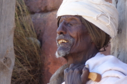 „Den Kopf leicht in den Nacken gekippt, saß er offnen Mundes da, ein Greis mit eckiger Nase und auch sonst markant knochigen Gesichtszügen, vielleicht ein Heiliger. Seine langen Fingernägel waren so stark ausgebleicht, daß sie wie weiß lackiert wirkten, sein schütterer Kinnbart war weiß, seine Zähne waren’s, der Kaftan, der Turban – so viel Weiß, daß seine tiefdunkelbraune Haut von innen zu glimmen schien. Mindestens ein Heiliger, dachte Trattner, ein Seher, der gar nicht mehr hinschauen muß, um zu erkennen.“