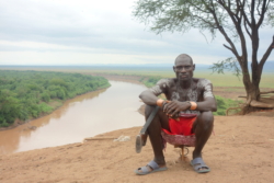 „Einer der Krieger saß etwas abseits der andern auf seinem winzigen Hocker, bekleidet mit Plastikschlappen, kurzen Hosen, Messingreifen an beiden Handgelenken und den Kabeln, die aus seinen Ohren zum Handy führten. Er sah dermaßen brutal und männlich aus, daß man Angst vor ihm bekommen konnte. Den Trageriemen seiner Kalaschnikow hatte er sich übern Oberschenkel gelegt, Schulterstück und Handlauf waren aus Holz. Er sah aus, als habe er unzählige Feinde auf dem Gewissen, an seinen Oberarmen war allerdings keine einzige Narbe zu sehen.“