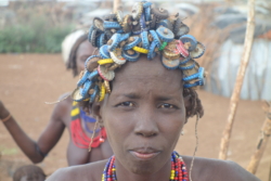 „Jede der Frauen hatte sich mit einem verschieden verrückten Kopfputz versehen, eine mit eine Haube aus Kronkorken, an einer Schnur aufgefädelt. Eine andre trug Ketten aus Kronkorken um beide Fußfesseln, die jüngeren zeigten ihre Brüste. Das sei alles nur Show, tat es Steve ab, Frauen mit irgendwelchen Perücken aus Kronkorken gebe’s in jedem Dorf. Auf diese Weise würden sie schnelles Geld zu verdienen, statt ihre Felder zu bestellen. Wenn genug Touristenbusse kämen, könnten sie sich von den Einnahmen nach drei Tagen eine Kuh kaufen.“